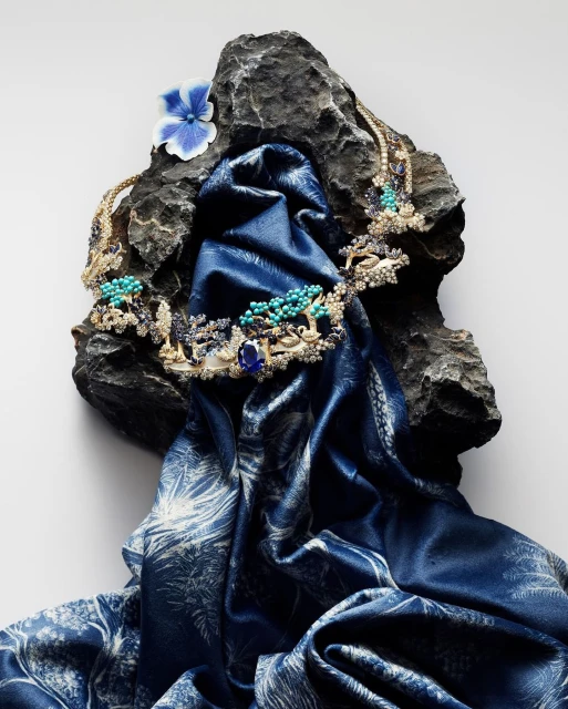 Elegant gold jewelry necklace with precious stones displayed on a dark rock, accented by a blue silk scarf and a delicate blue flower.