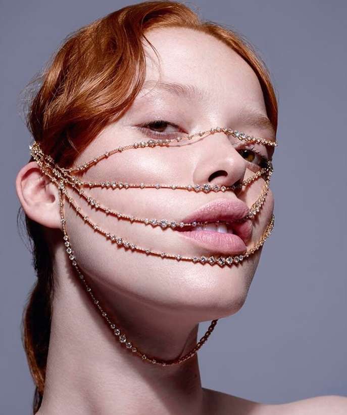 Portrait of an adult female with red hair wearing a gold colored beaded jewelry face covering against a gray backdrop in a studio setting.
