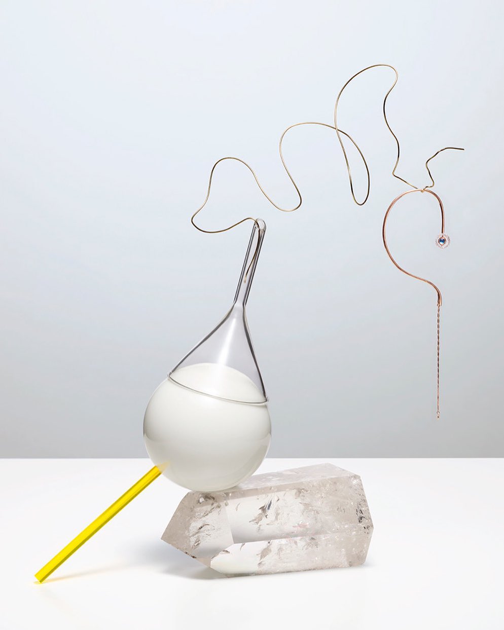 Still life shot of a glass flask containing a white sphere, balanced on a clear quartz crystal with gold wire and a yellow straw.