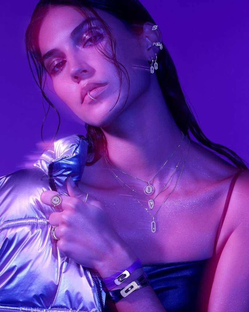 Portrait of a young adult female with dark hair and jewelry, wearing a puffer jacket in a neon violet and blue light setting.
