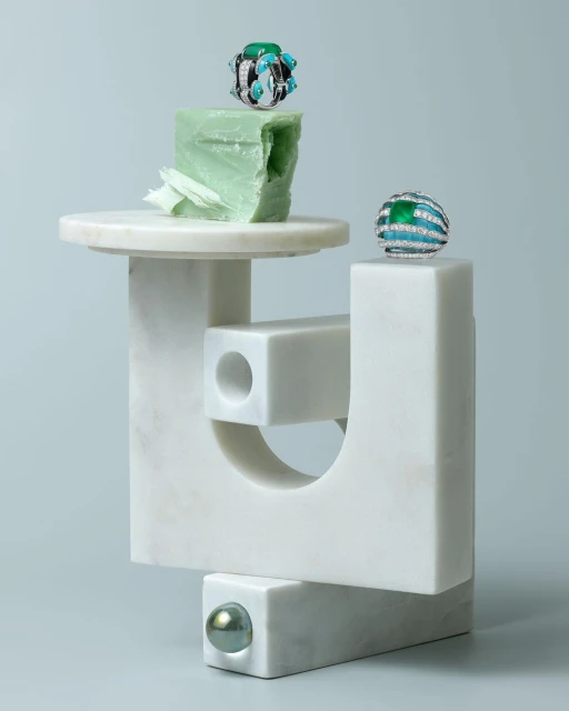 Close-up still life of two gemstone rings displayed on an abstract sculpture with geometric shapes in a softly lit, studio setting.