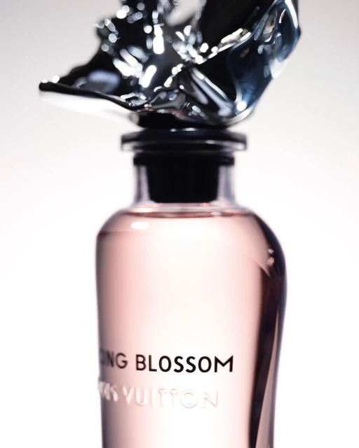 Close-up of a Louis Vuitton perfume bottle with a silver flower-shaped cap, set against a bright white background. Elegant fragrance shot.