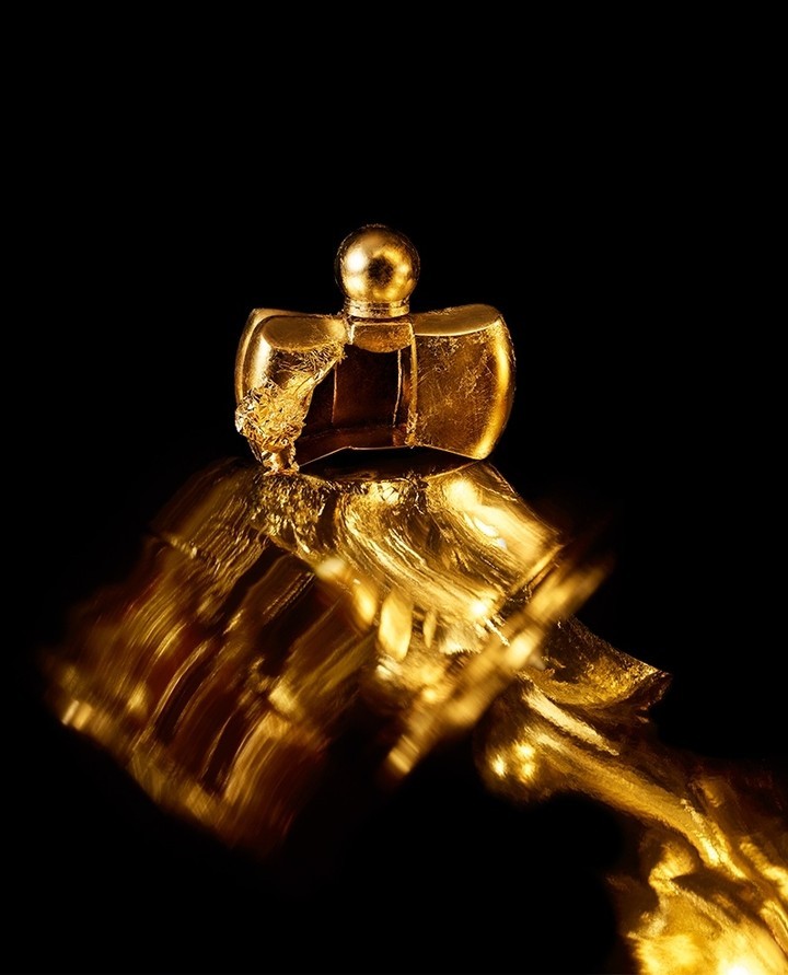 Close-up studio shot of a textured, partially broken golden perfume bottle against a black background, showcasing light and reflection.