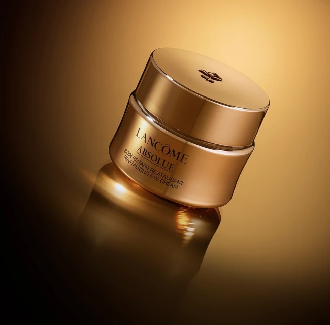 Close-up studio shot of Lancôme Absolue Revitalizing Eye Cream, featuring its golden container and lid, against a matching gold gradient backdrop.