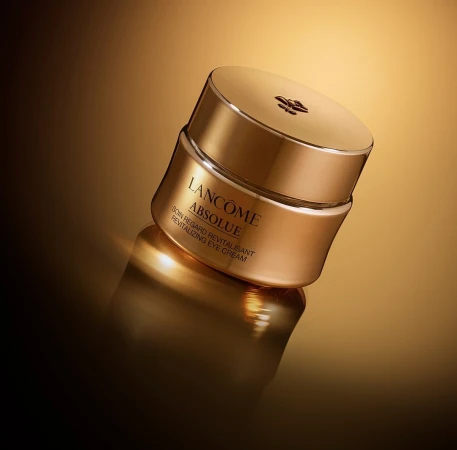 Close-up studio shot of Lancôme Absolue Revitalizing Eye Cream, featuring its golden container and lid, against a matching gold gradient backdrop.