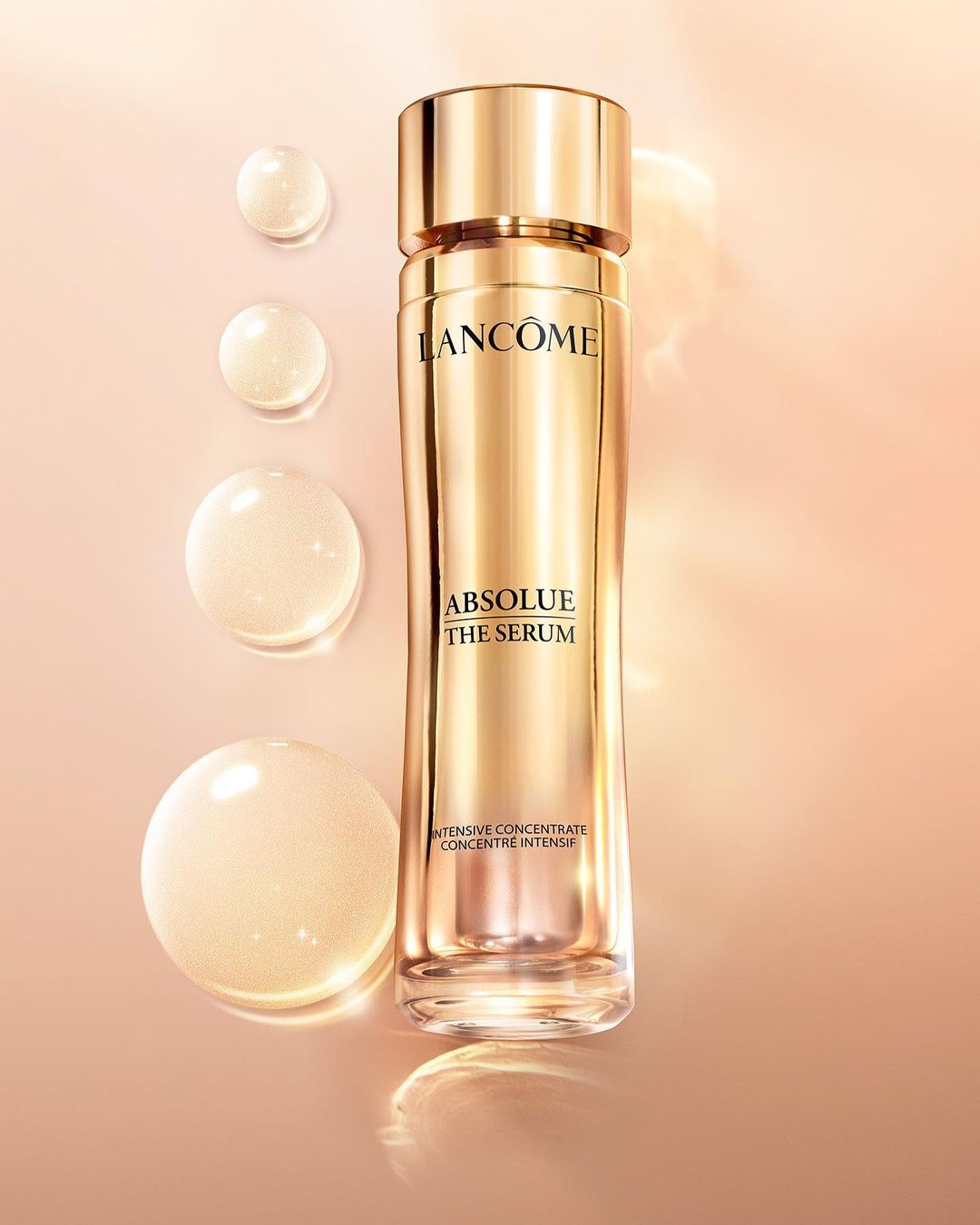 Gold Lancôme Absolue The Serum bottle with water droplets against a soft peach background, highlighting luxury skincare.