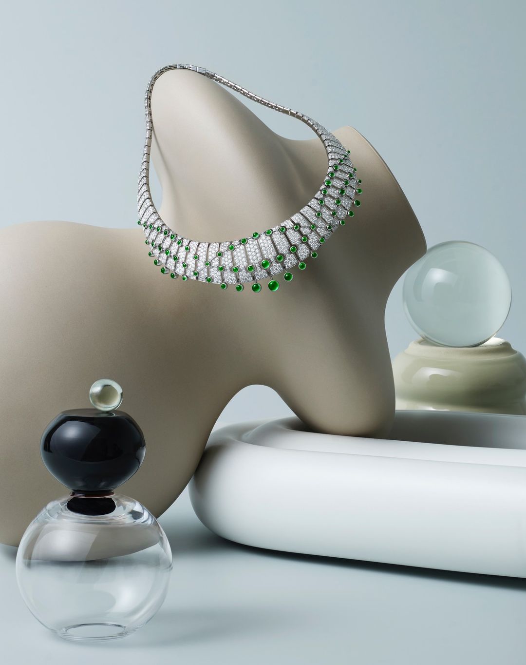 Elegant diamond and emerald necklace displayed on a sculptural form with perfume bottle and glass ball against light blue background.