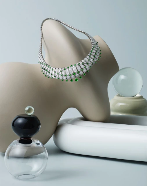 Elegant diamond and emerald necklace displayed on a sculptural form with perfume bottle and glass ball against light blue background.