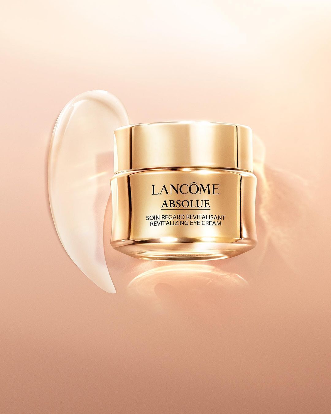 Lancôme Absolue revitalizing eye cream jar with golden lid next to cream smear on warm-toned background. Beauty shot.