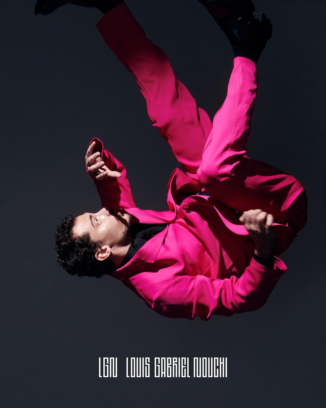 Man in bright pink suit with dark shoes poses at an angle against a dark grey background in a fashion photography studio shoot.