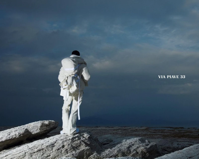 Back view of a person in white outerwear, wearing a backpack, standing on rocky terrain beneath a cloudy sky, Via Piave 33 text.