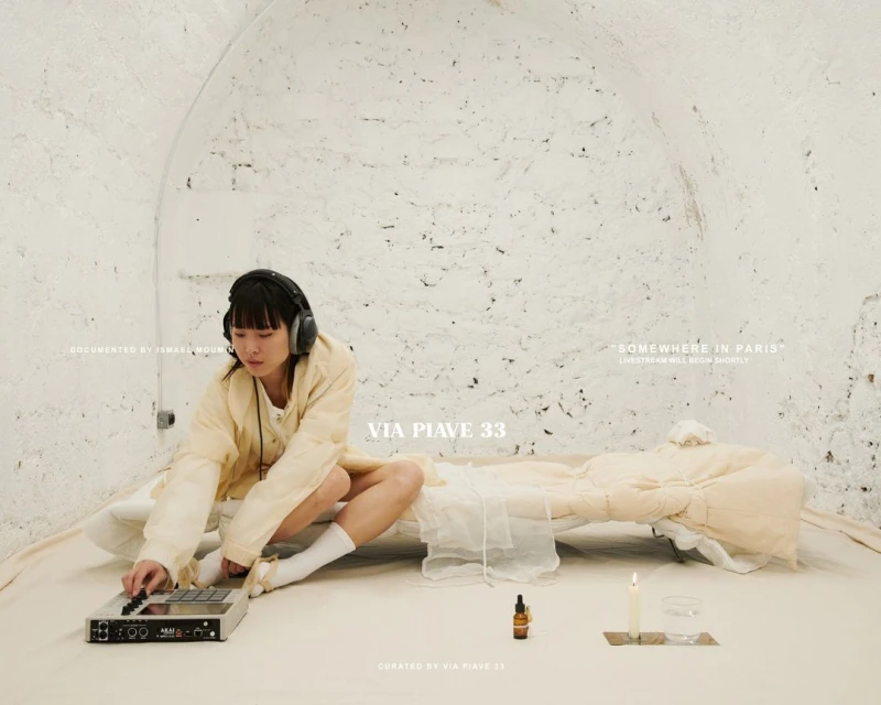 Woman sits on the floor in a studio-like space, wearing headphones and operating a beat machine. A candle, bottle, and water glass rest nearby.
