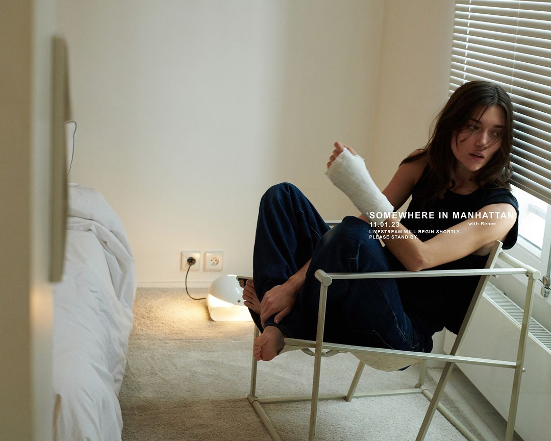 A woman with a cast sits in a chair near a window in a room with minimalist decor, illuminated by a modern lamp and natural light.
