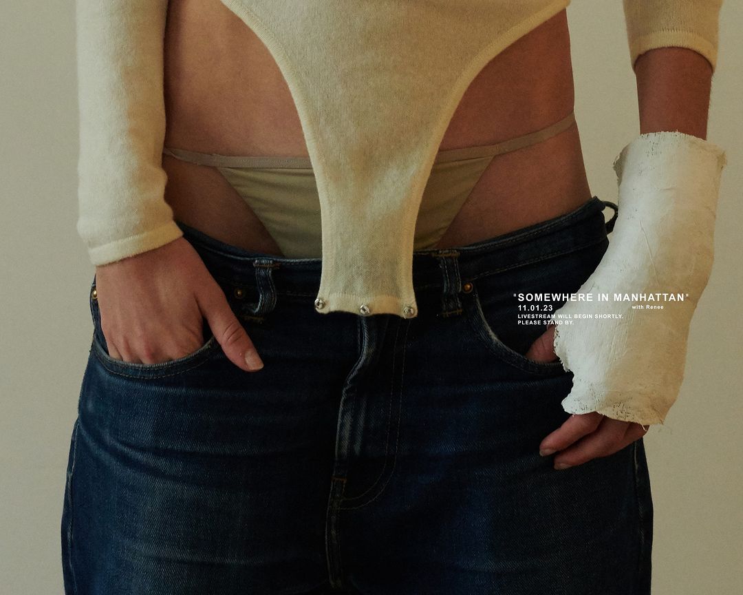 A slender, light-skinned person stands indoors wearing a cream-colored long-sleeved bodysuit, blue jeans, and a white cast on their right wrist.