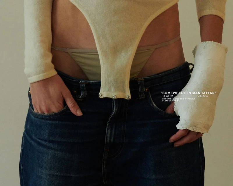 A slender, light-skinned person stands indoors wearing a cream-colored long-sleeved bodysuit, blue jeans, and a white cast on their right wrist.