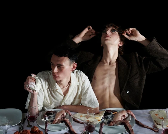 Two young men seated at a table with unconventional meal featuring plates with meat, against a stark black background.