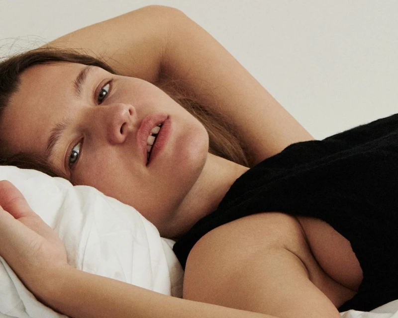 Close-up portrait of a woman in a black top, lying on a white pillow, looking at the camera with a neutral expression in indoor setting.