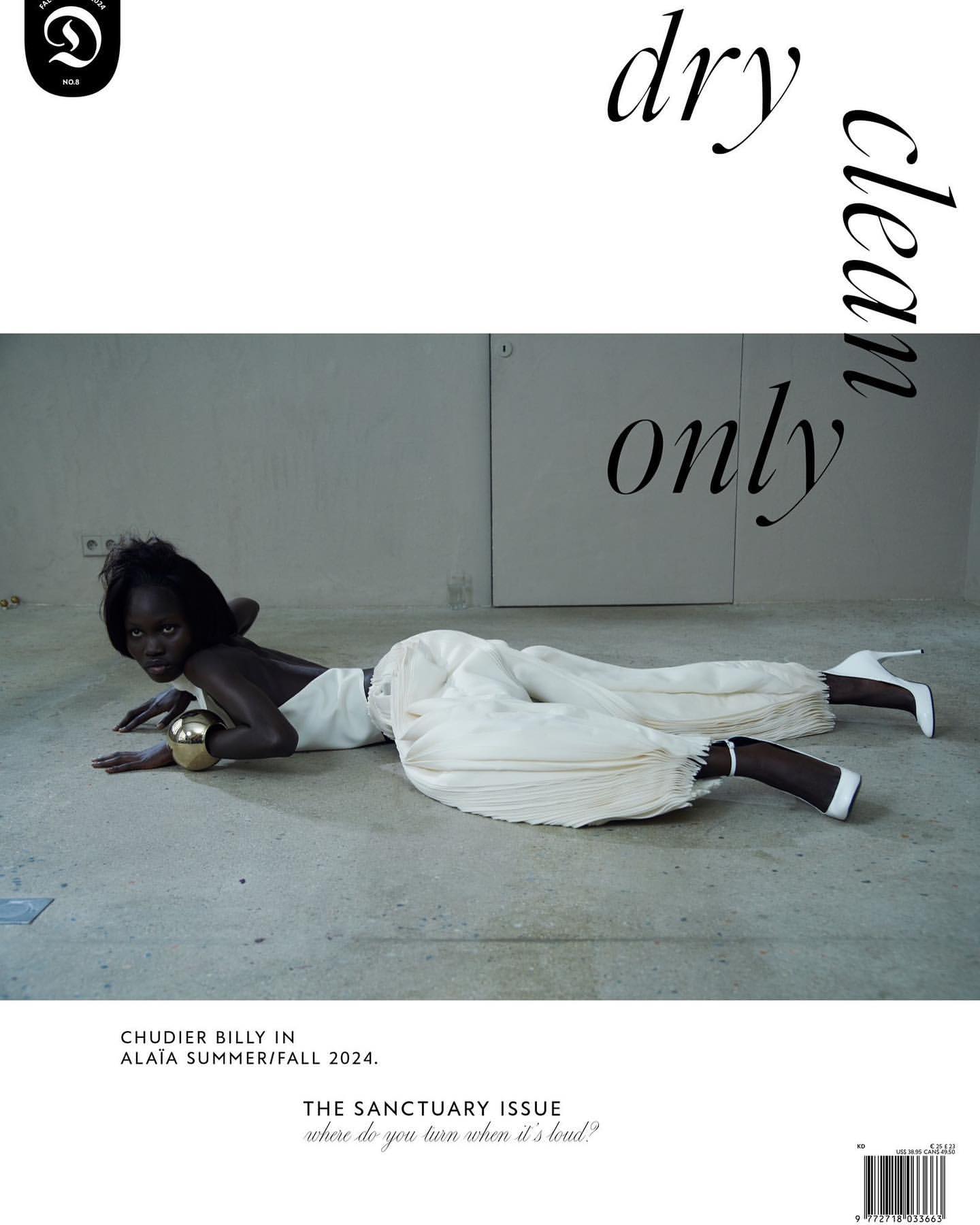 Editorial fashion shot of a Black female fashion model reclining in white pleather pants and heels with a gold arm cuff on a gray floor.