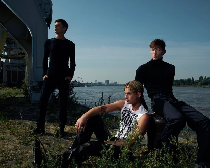 Three young adult men pose outdoors in a fashion editorial on an overcast day near a body of water with an urban cityscape in the background.
