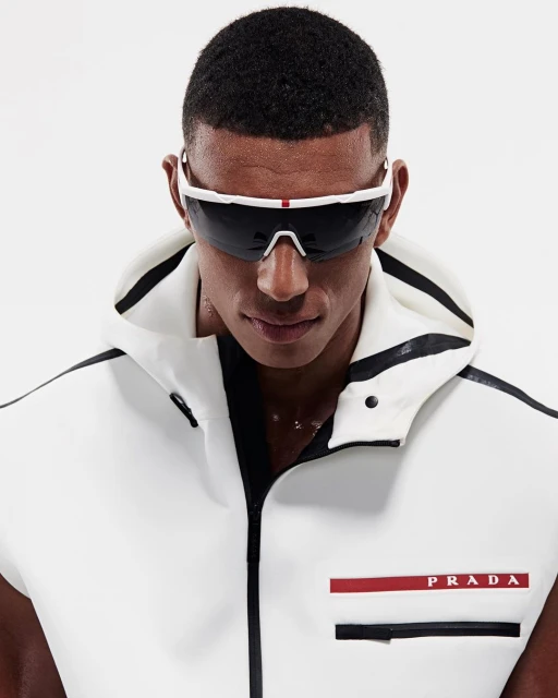 Adult male poses indoors wearing a sleeveless white hooded jacket and sunglasses; the jacket features a Prada logo.