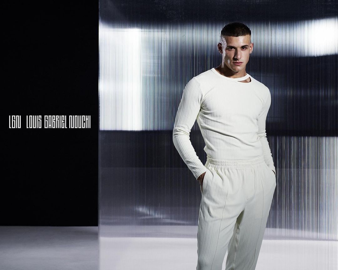Studio shot of a young adult male model in an off-white ribbed long-sleeve shirt and pants, posing in front of a silver corrugated background.