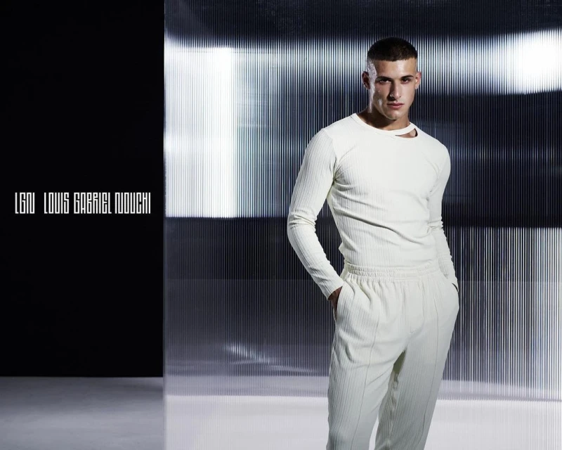 Studio shot of a young adult male model in an off-white ribbed long-sleeve shirt and pants, posing in front of a silver corrugated background.