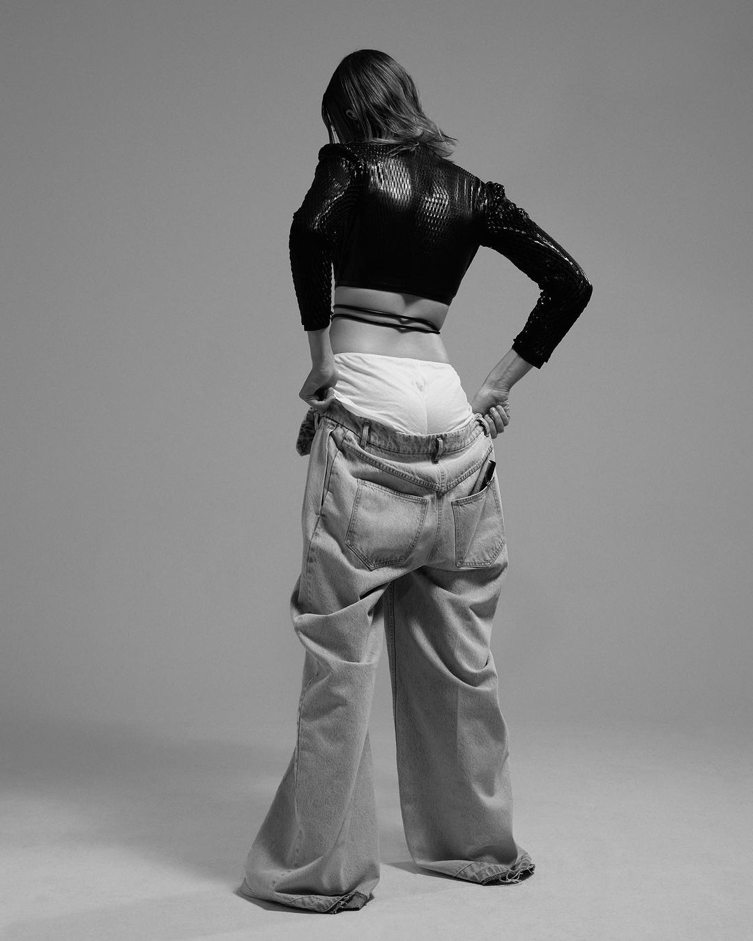 A woman with short brown hair, from the rear, poses in a black sequined top and baggy jeans in a studio with a gray background.