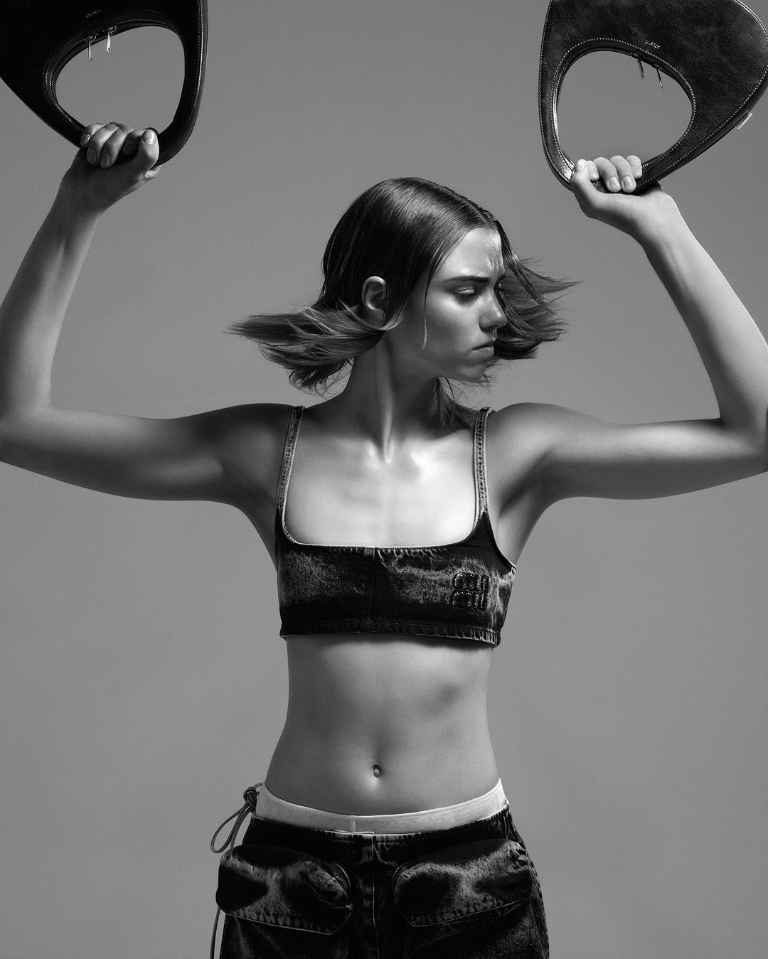 Monochrome portrait of a young woman in denim crop top and low-rise jeans, holding handbag handles overhead in a studio shot.