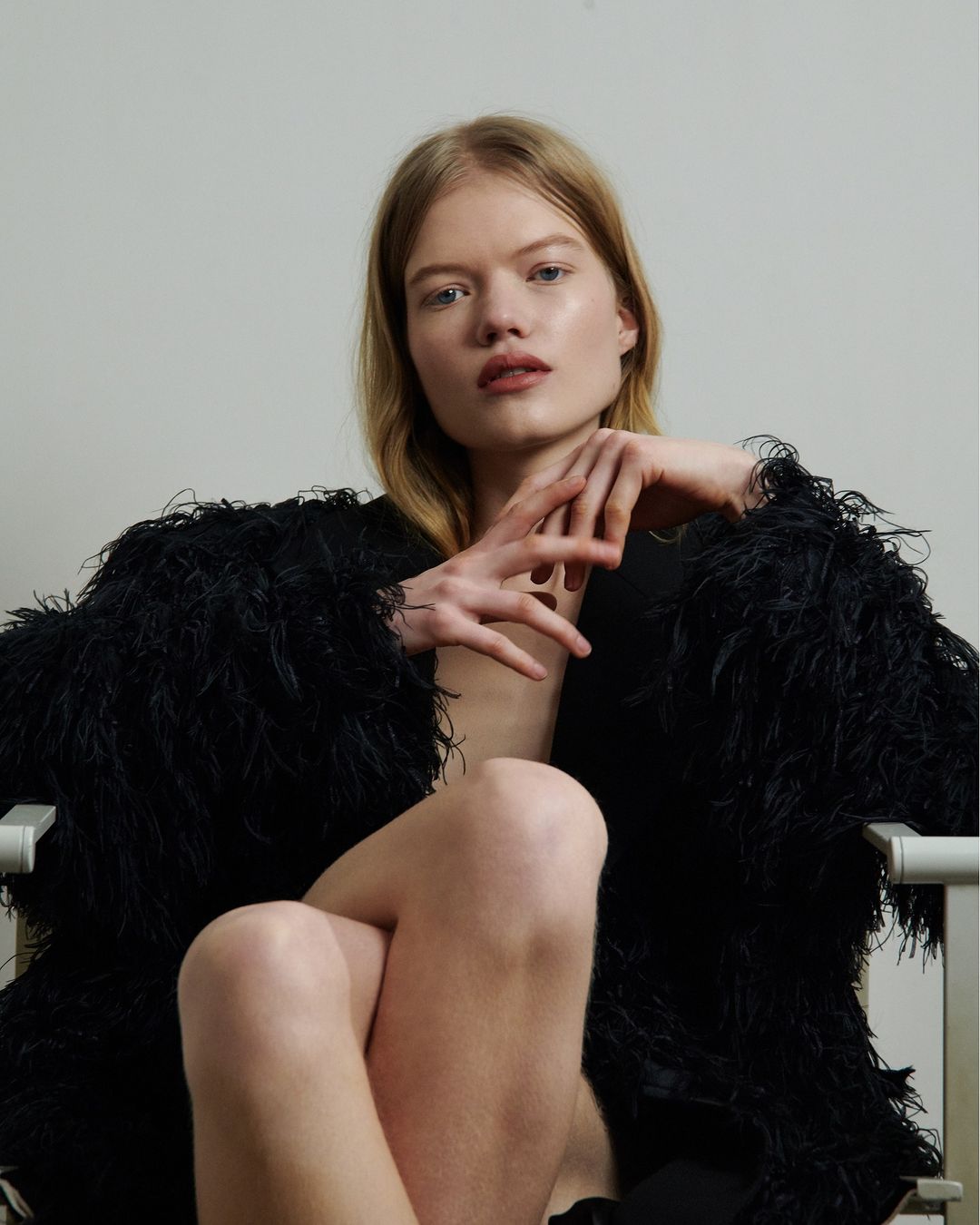 Portrait of a fair-skinned blonde woman wearing a black feathered jacket, seated with her legs crossed in a light indoor setting.