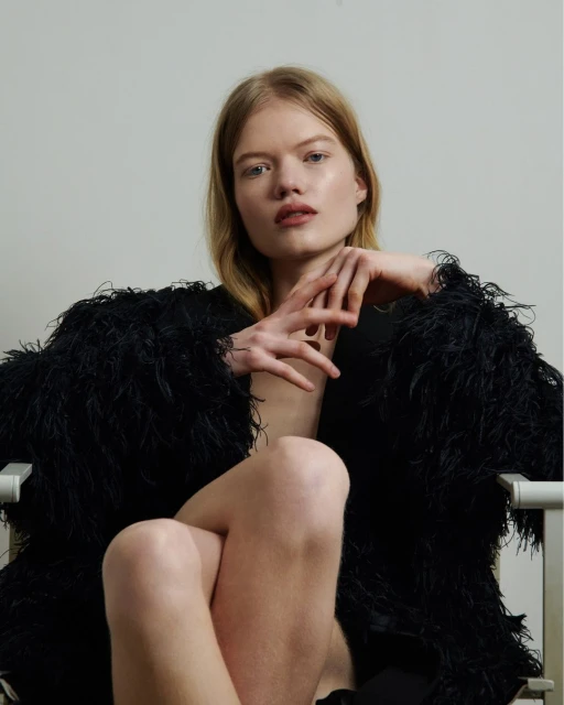 Portrait of a fair-skinned blonde woman wearing a black feathered jacket, seated with her legs crossed in a light indoor setting.