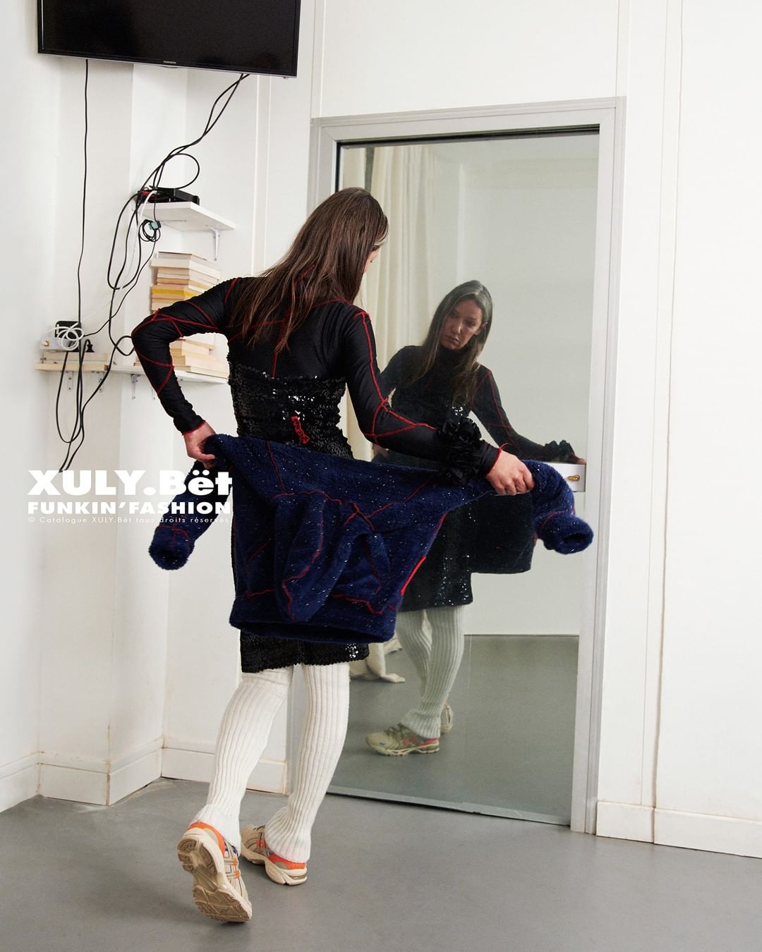 Woman in black outfit checks her reflection in an indoor setting with white walls and a TV, holding a navy jacket with red accents.