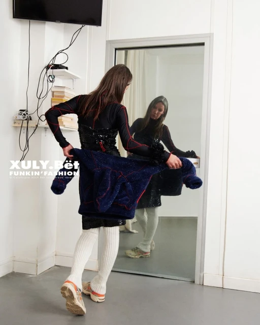 Woman in black outfit checks her reflection in an indoor setting with white walls and a TV, holding a navy jacket with red accents.