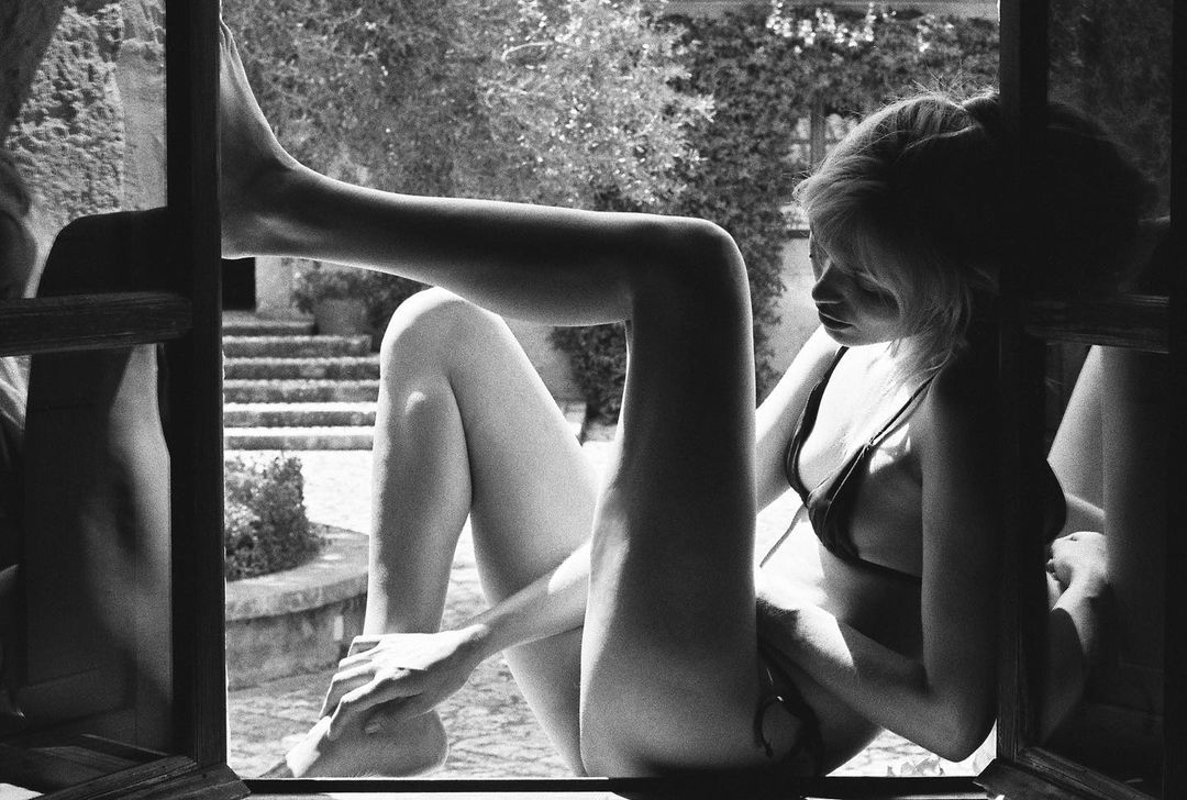Monochrome image of an adult female in lingerie posing in a window with a natural outdoor background. Black and white photography.