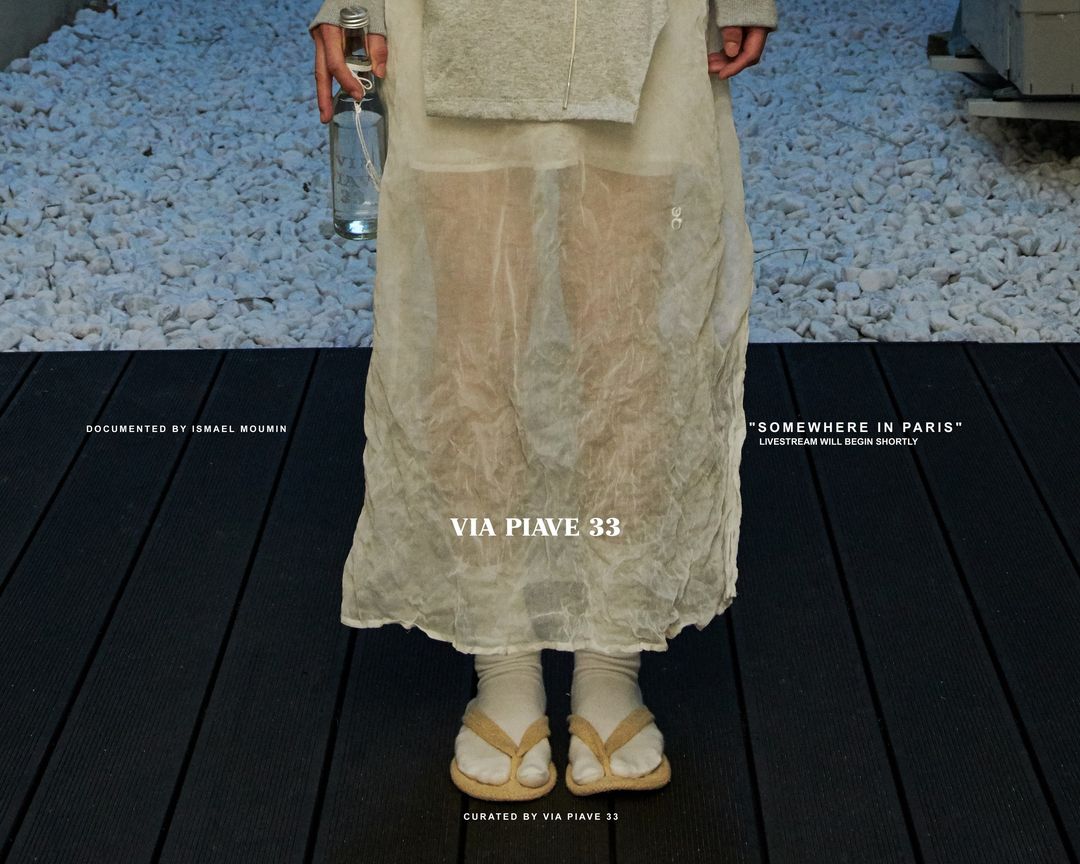 Woman in translucent dress and flip-flops stands on black planks with a bottle. White rocks in the background. Fashion editorial.