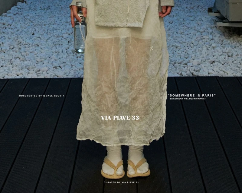 Woman in translucent dress and flip-flops stands on black planks with a bottle. White rocks in the background. Fashion editorial.
