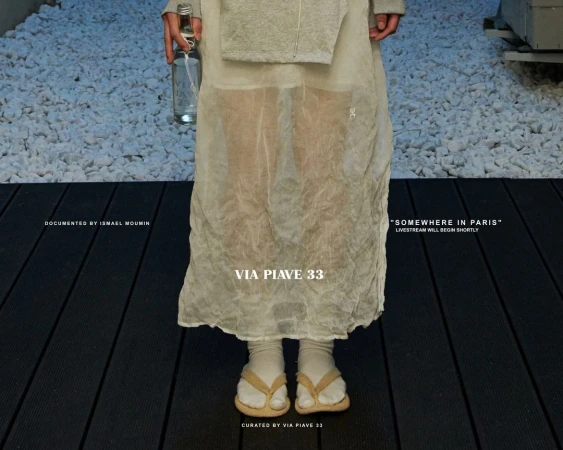 Woman in translucent dress and flip-flops stands on black planks with a bottle. White rocks in the background. Fashion editorial.