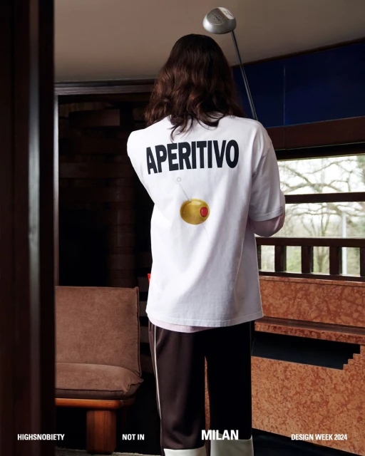 Back view of a person in a white t-shirt with 'Aperitivo' text and an olive graphic, standing in a naturally lit interior space.