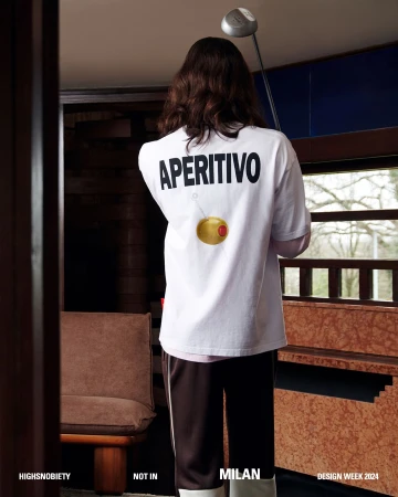 Back view of a person in a white t-shirt with 'Aperitivo' text and an olive graphic, standing in a naturally lit interior space.