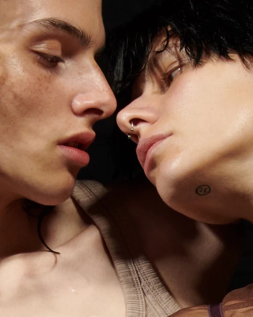 Close-up of two faces in soft lighting, showcasing smooth skin, nose piercings, and neutral expressions, set against a dark background.