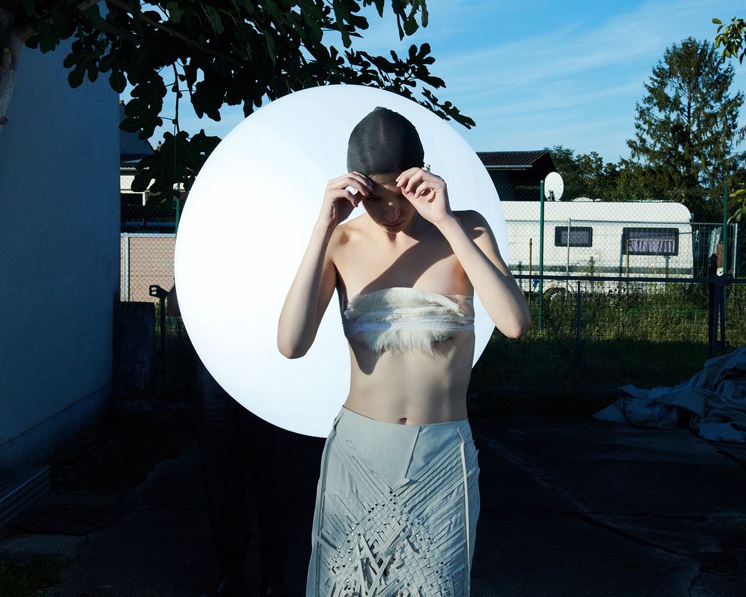 A young woman wearing a bandeau top and skirt poses outdoors with a large round light and camper in the background.