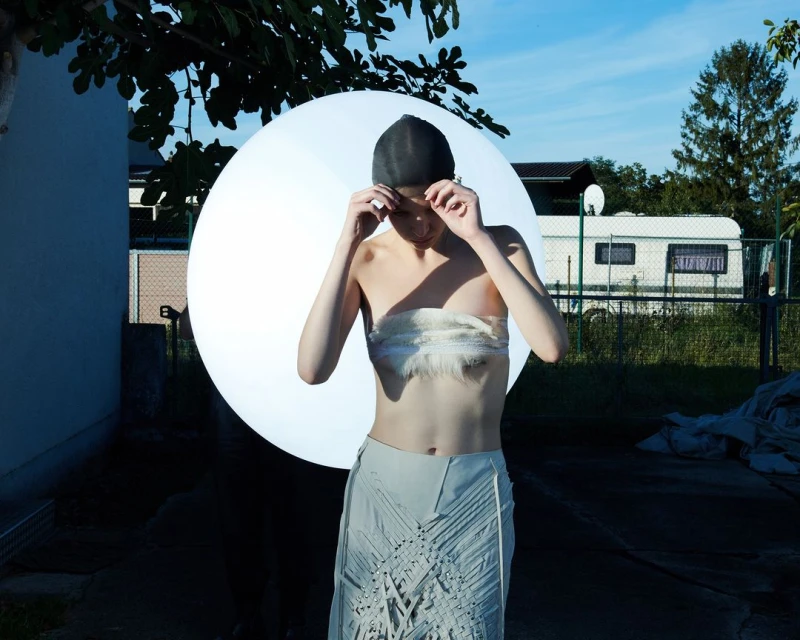 A young woman wearing a bandeau top and skirt poses outdoors with a large round light and camper in the background.