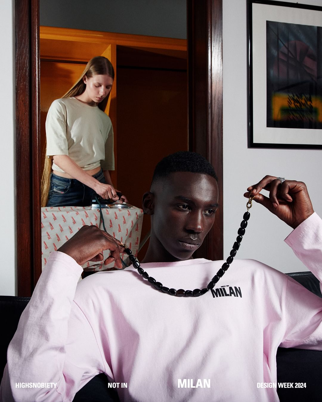 A black model in a pink shirt with a black necklace is photographed in an indoor setting, with a woman ironing in the background.
