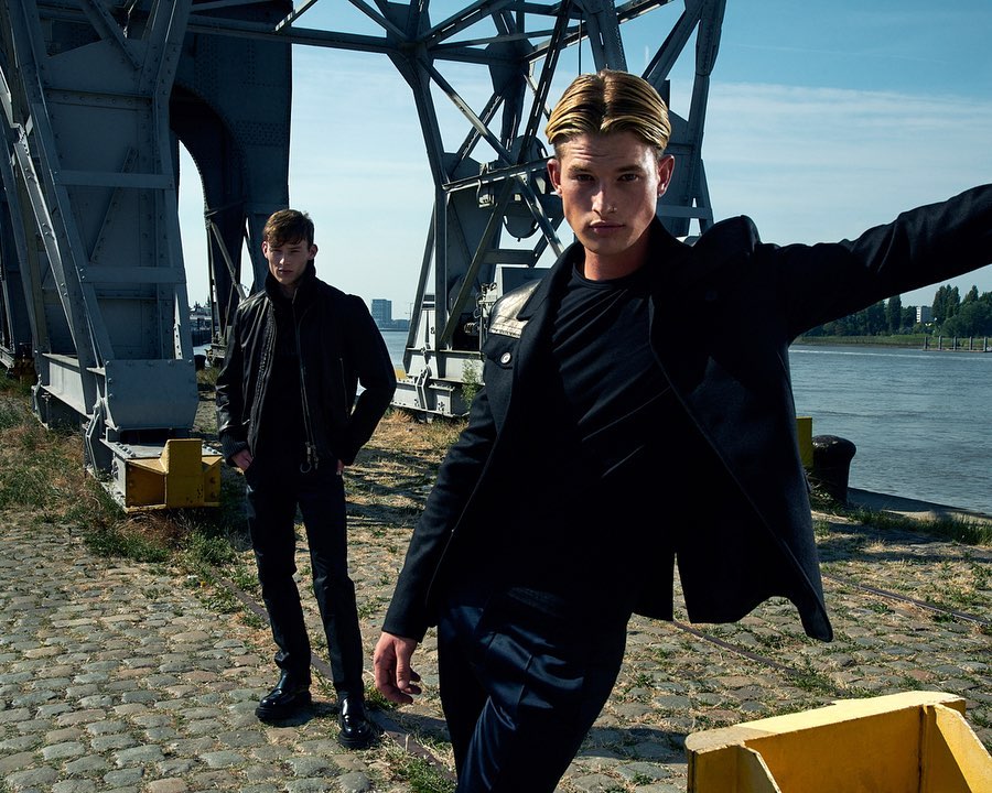 Two adult men pose outdoors near industrial structures and a body of water, wearing dark jackets and pants in a fashion editorial setting.