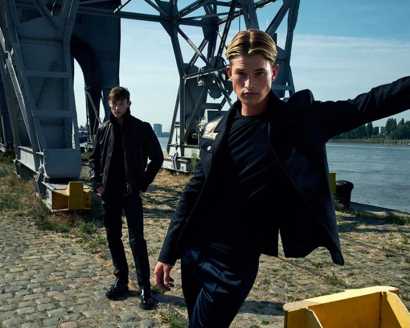 Two adult men pose outdoors near industrial structures and a body of water, wearing dark jackets and pants in a fashion editorial setting.
