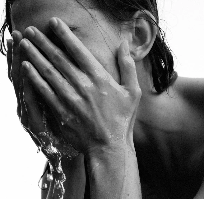Close-up black and white shot of an adult with water running down their face and hands, covering their face against a white backdrop.