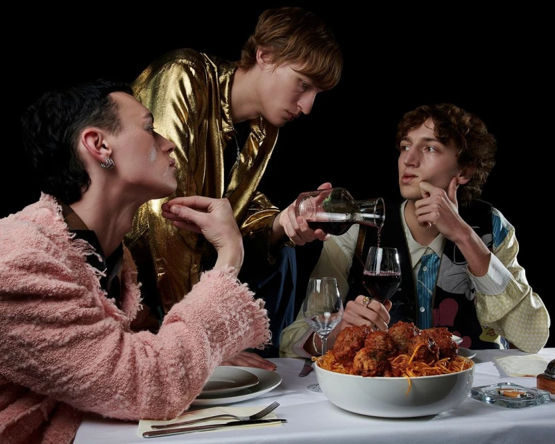 Three stylish people at dinner: one pours wine, another is in a pink coat, and a large bowl of spaghetti is in the foreground.