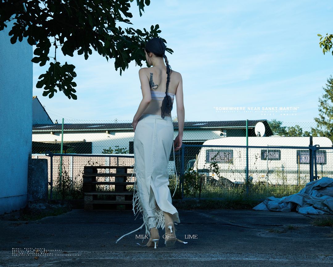 A slender woman with a braid stands in the shade on a sunny day wearing a hat, strapless top, flowing skirt, and kitten heels.
