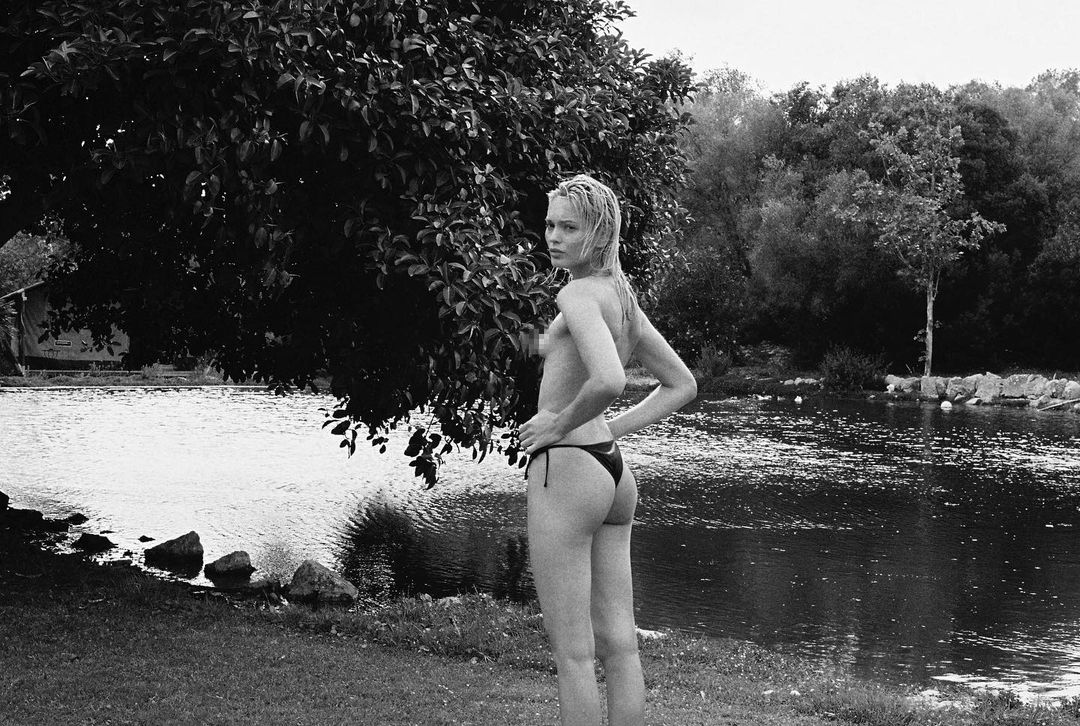 Woman in bikini poses lakeside next to leafy tree in black and white photograph featuring natural outdoor environment with ripples.