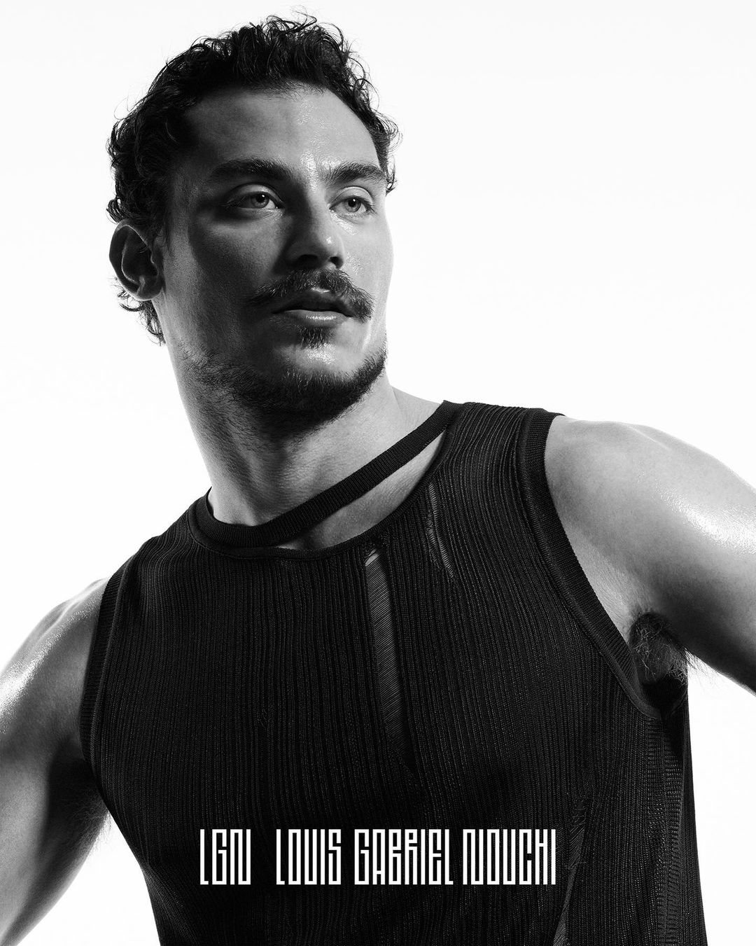 Monochrome portrait of a man in a black tank top looking away, posing against a bright white background in a studio environment.