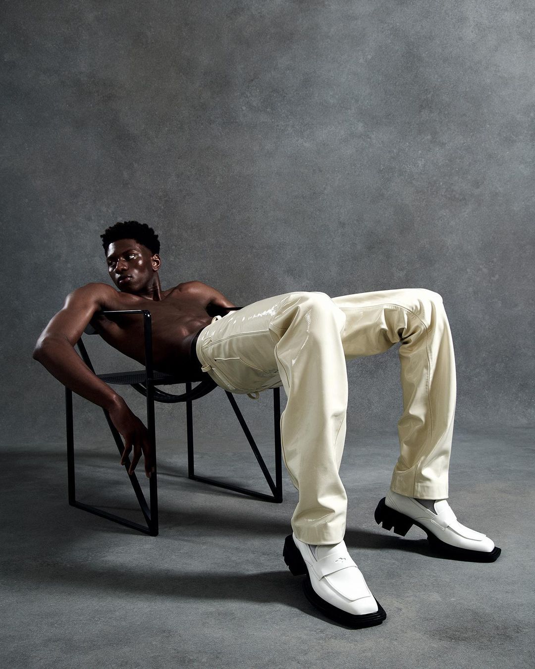 A shirtless Black male fashion model poses sitting in a chair wearing glossy off-white pants and white penny loafers against a grey backdrop.